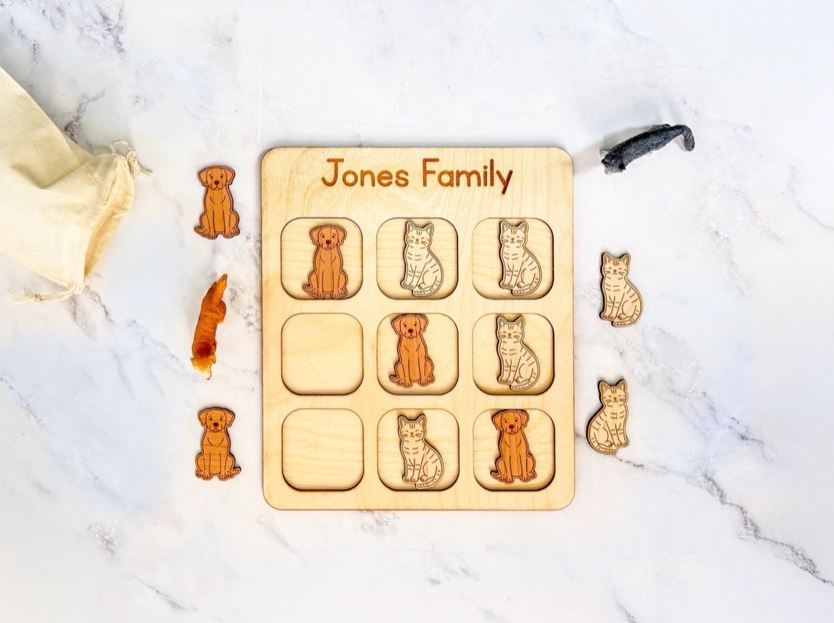 Personalized wooden tic tac toe board engraved 'Jones Family' with laser-engraved dog and cat playing pieces mid-game on the three-by-three grid, extra pieces, animal figurines, and a muslin bag on white marble