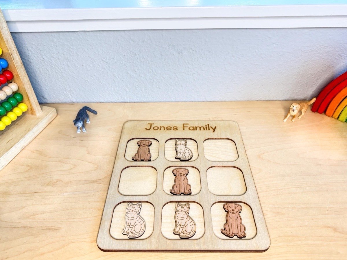 Cats and dogs tic tac toe board on a playroom desk between a bead abacus, rainbow stacker, and toy animal figurines, six wooden pieces mid-game on the personalized grid