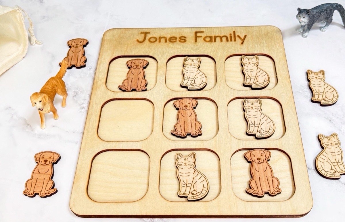 Angled view of a cats versus dogs tic tac toe game in progress on a board engraved 'Jones Family,' toy cat and dog figurines and extra wooden pieces around the marble surface