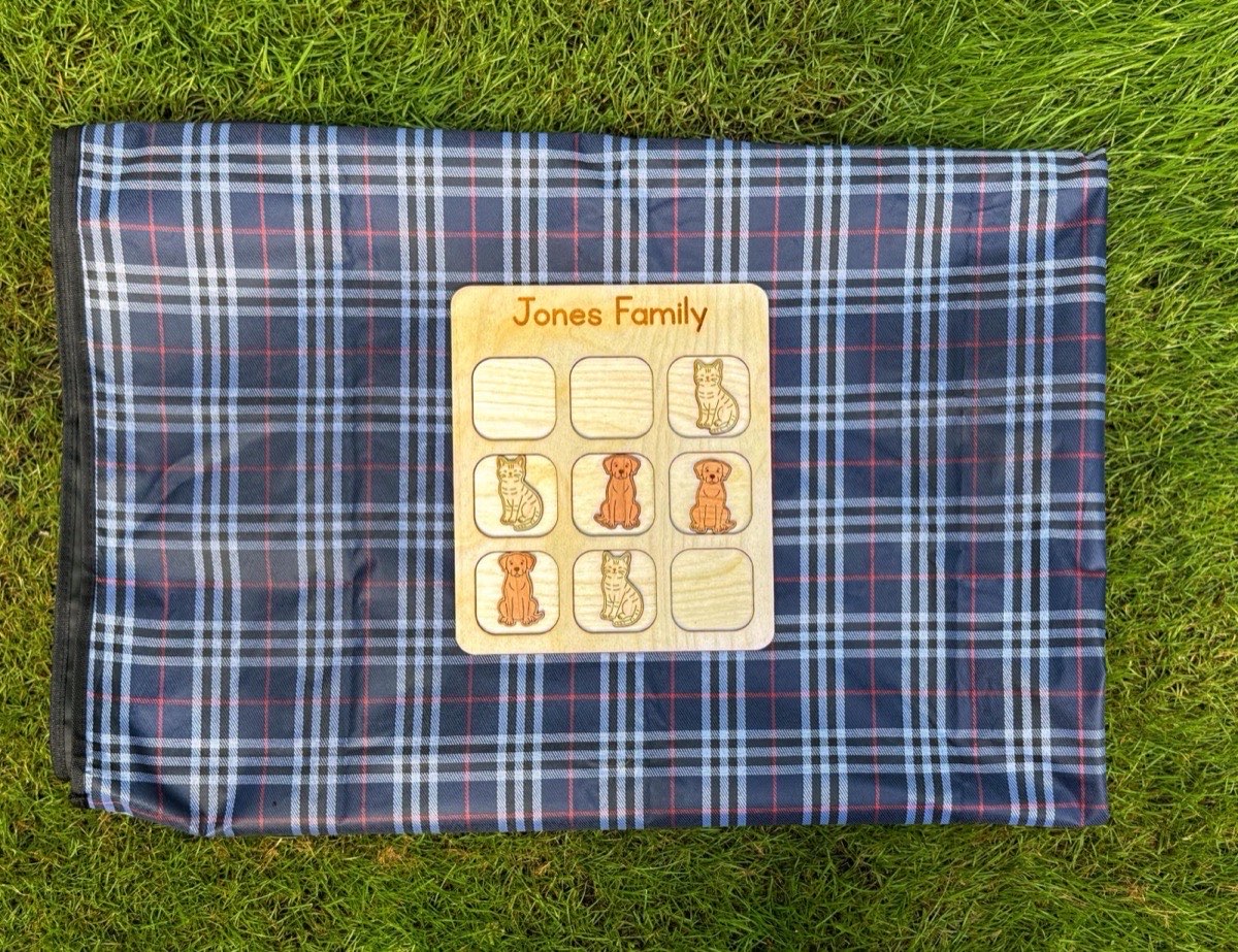 Wooden family tic tac toe board on a navy plaid picnic blanket in grass, dog and cat game pieces placed across the grid for a backyard match