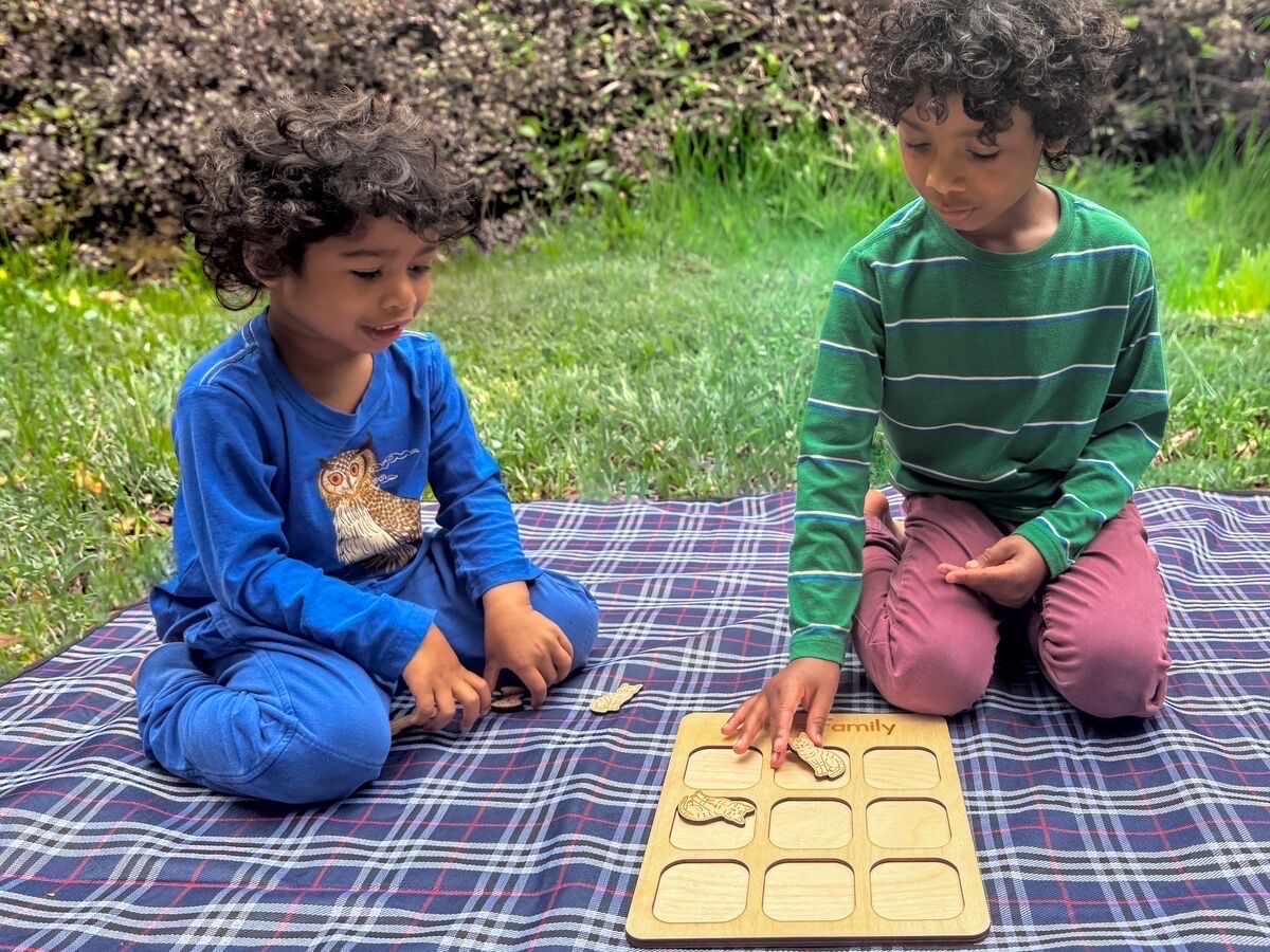 Two young children playing wooden cat and dog tic tac toe outdoors on a plaid picnic blanket, setting laser-engraved cat pieces into a neutral Baltic birch board with a personalized family name engraved across the top