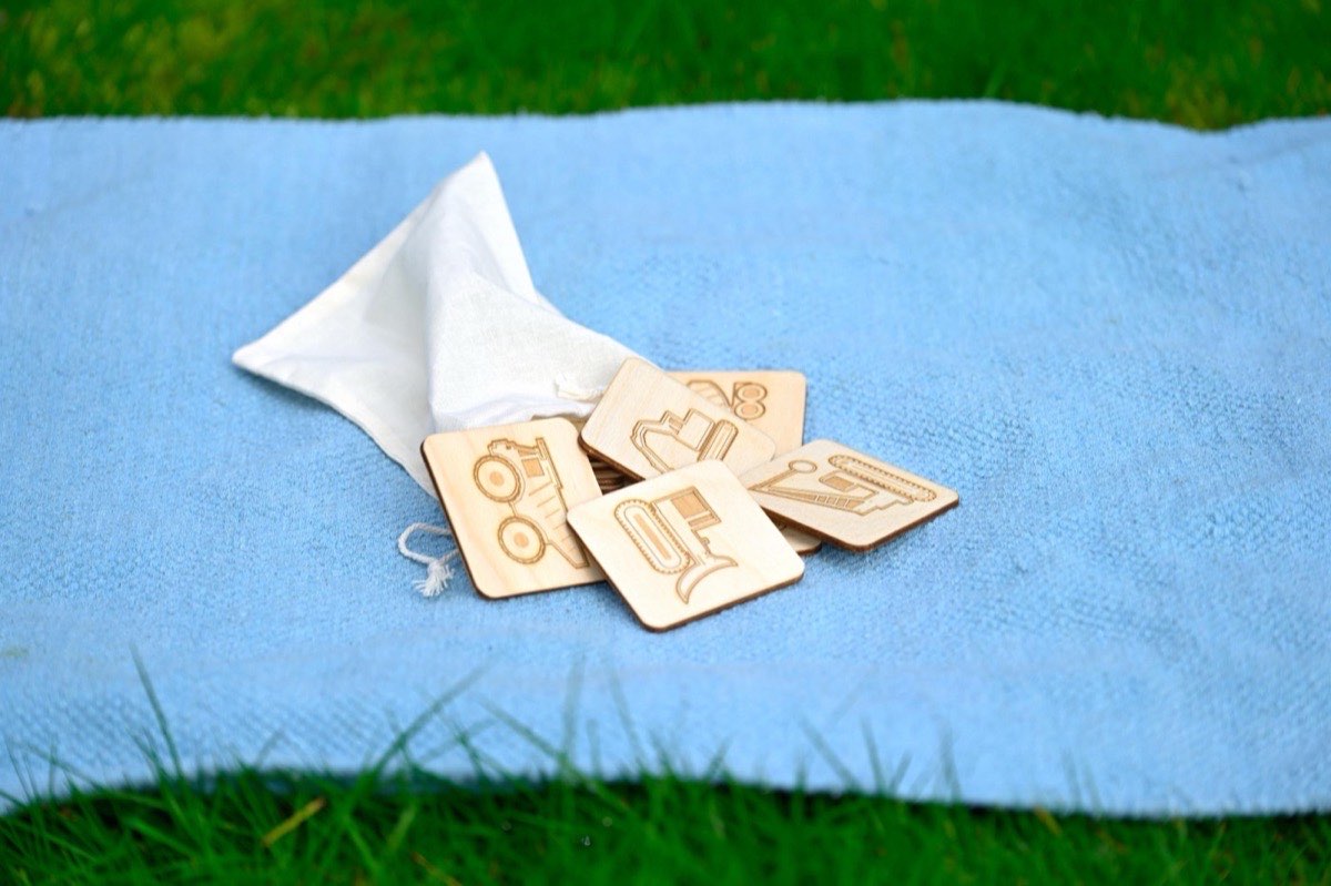 Construction vehicle memory match tiles spilling out of a cotton muslin drawstring bag onto a blue blanket in the grass
