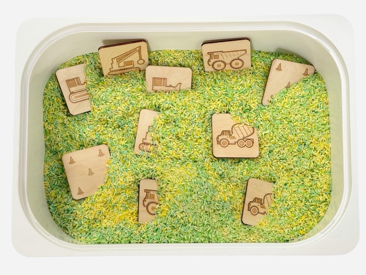10-piece wooden construction vehicle memory match set in neutral Baltic birch scattered across a green and yellow rice sensory bin, featuring laser-engraved dump truck, excavator, cement mixer, bulldozer, crane truck pairs