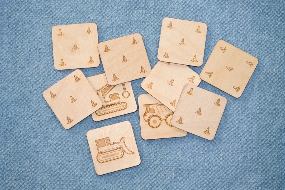 Ten neutral-colored wooden construction vehicle memory match tiles scattered on a blue blanket, showing both the laser-engraved vehicle fronts (crane, dump truck, bulldozer) and the traffic cone pattern on neutral Baltic birch backs