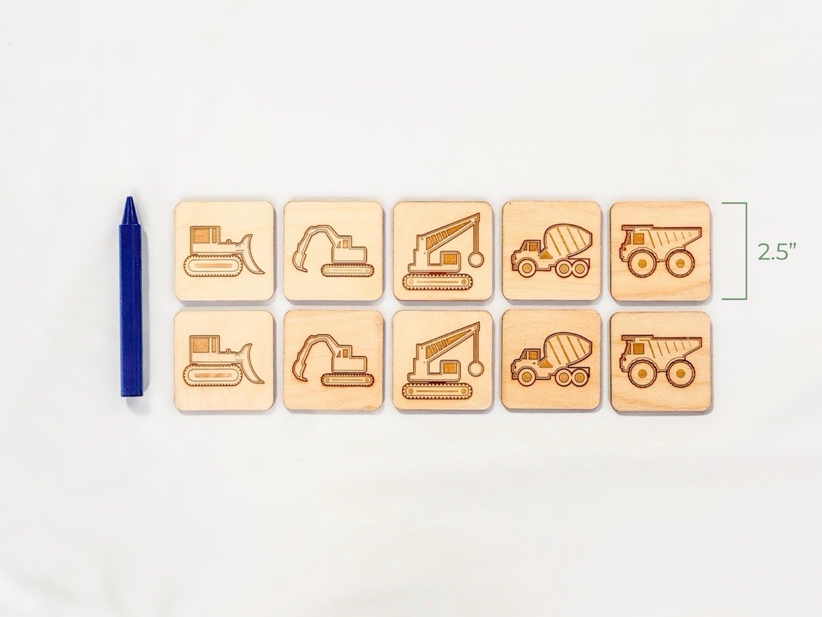 Ten-piece wooden construction vehicle memory match game shown beside a blue jumbo crayon for size reference, with five laser-engraved pairs of bulldozer, excavator, wrecking ball crane, cement mixer, and dump truck on neutral Baltic birch.  Pieces are 2.5" tall with rounded corners.  On a white fabric background