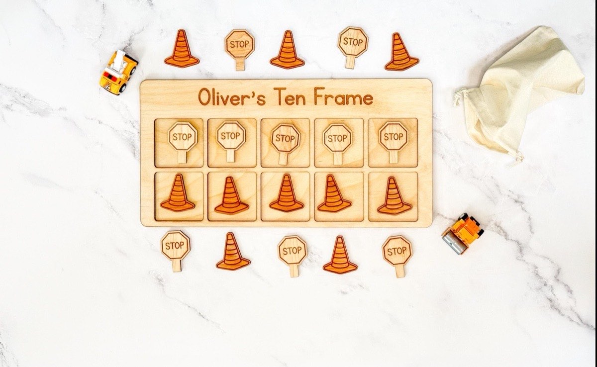 Personalized construction ten frame engraved 'Oliver's Ten Frame' on marble, five stop sign counters in the top row and five striped traffic cone counters below, toy work trucks and a muslin drawstring bag nearby
