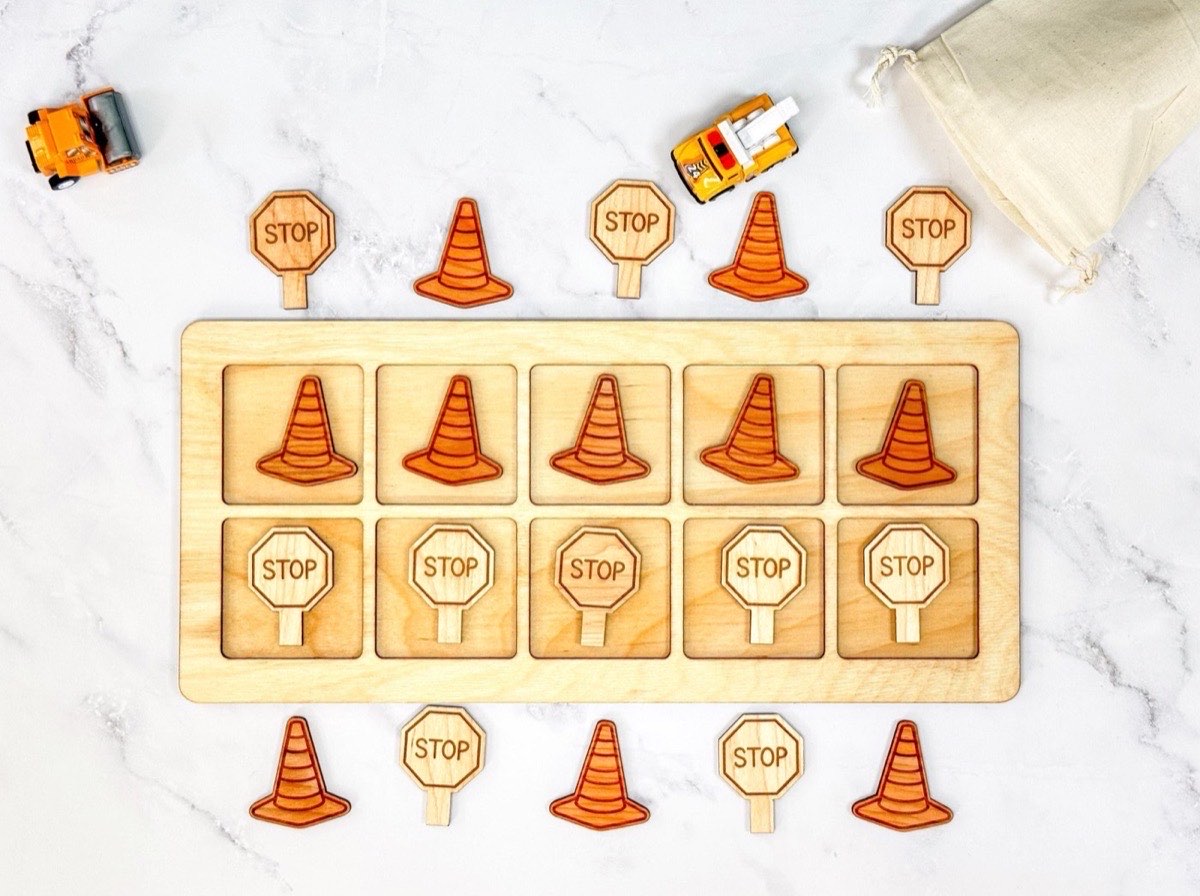 Non-personalized wooden ten frame with five traffic cones across the top row and five stop signs below, extra construction counters, toy trucks, and a natural muslin bag arranged around the neutral Baltic birch board