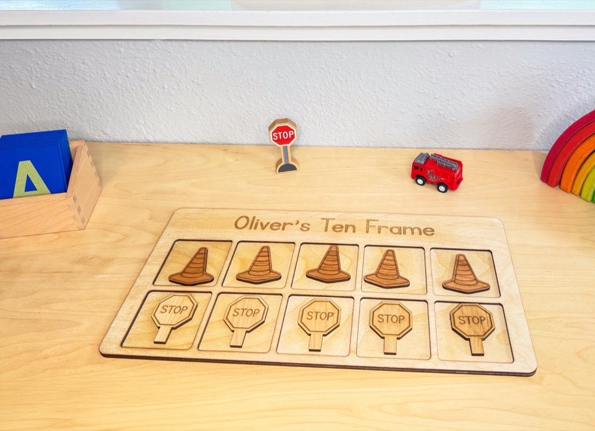 Construction ten frame on a playroom desk with a red toy fire truck, wooden stop sign, and rainbow stacker, five cone counters in the top row and five stop sign counters below