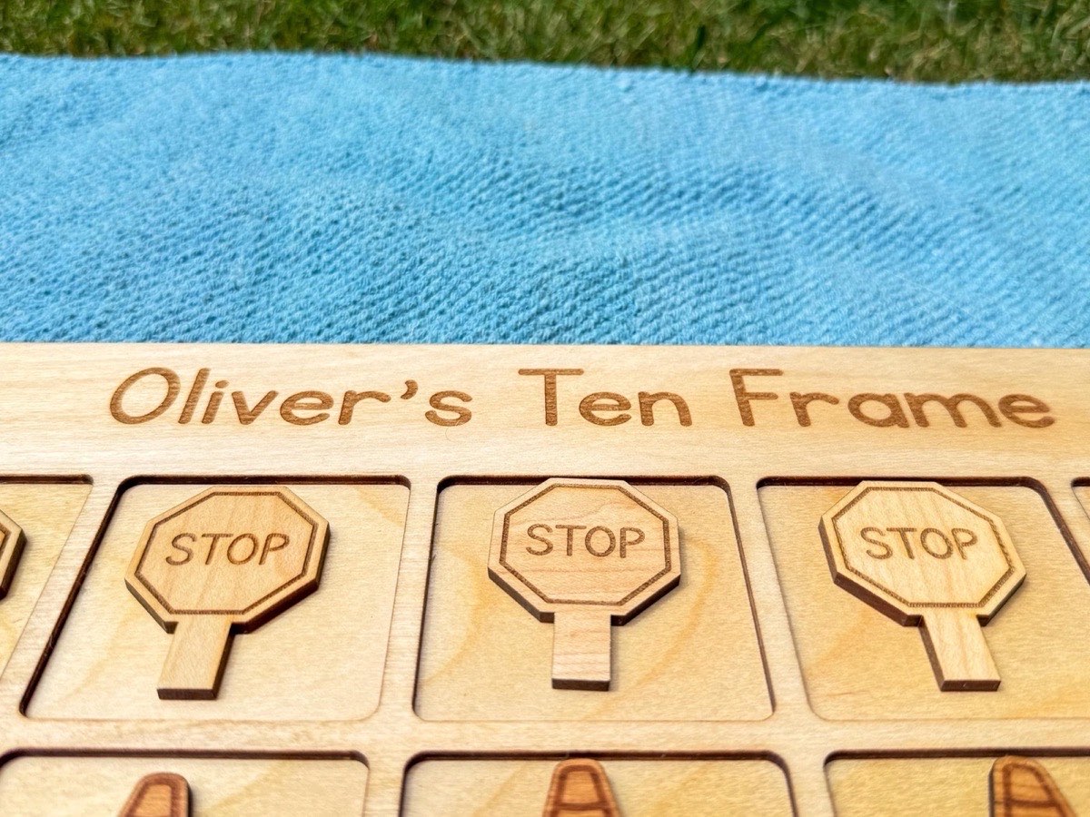 Close-up of the engraved 'Oliver's Ten Frame' lettering and three stop sign counters, showing laser-engraved detail and natural grain of the Baltic birch board on a blue blanket outdoors