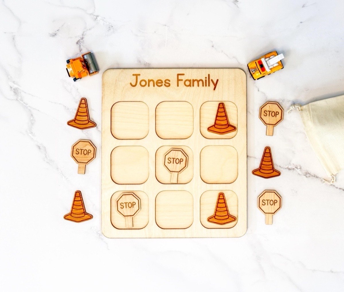 Personalized construction tic tac toe board engraved 'Jones Family' with traffic cone and stop sign playing pieces mid-game, toy work trucks and a muslin drawstring bag on the white marble surface