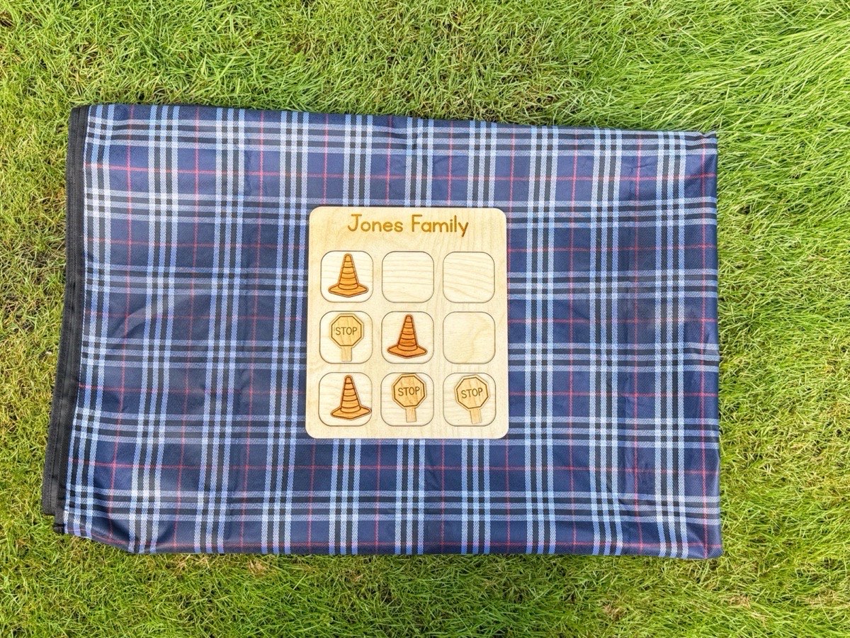 Construction tic tac toe board on a navy plaid blanket in grass, wooden cone and stop sign pieces placed across the family-name grid for backyard play