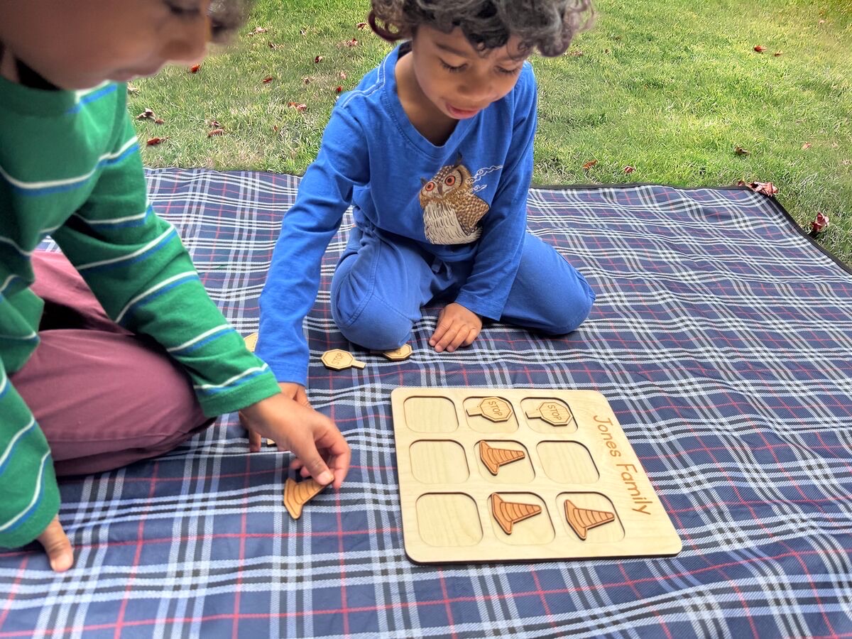 Children playing wooden construction tic tac toe on a picnic blanket in the grass, with laser-engraved orange traffic cone and stop sign pieces on a neutral Baltic birch board personalized Jones Family