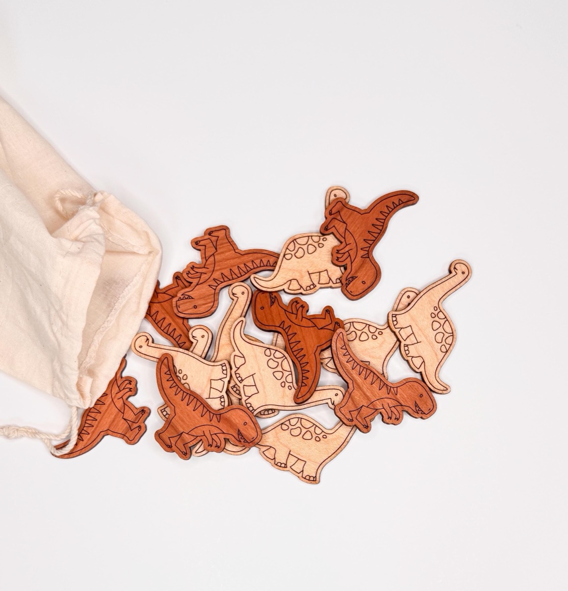 Wooden dinosaur manipulatives tumbling from a cotton drawstring storage bag, mixing T-Rex and brachiosaurus shapes in two natural wood tones for open-ended counting, sorting, and small world play