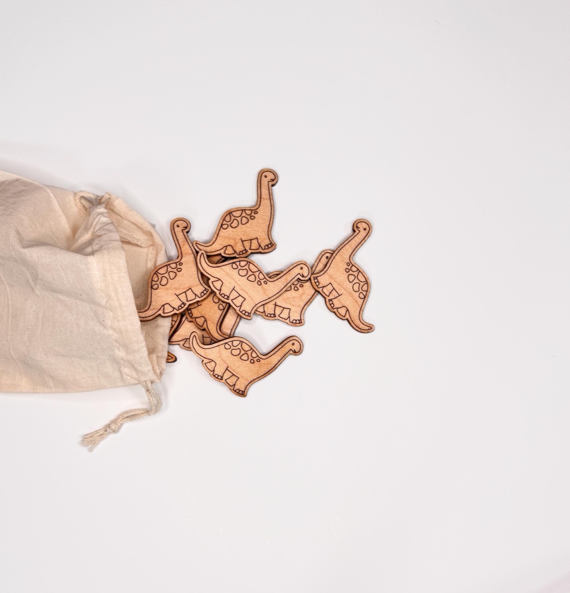 Brachiosaurus wooden counters in warm neutral tones spilling from a natural cotton drawstring bag, each engraved with a long curved neck, spotted back, and sturdy legs