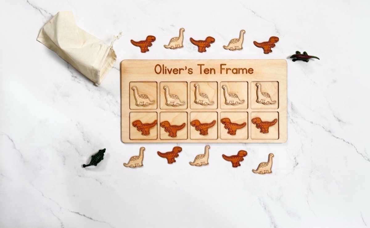 Personalized dinosaur ten frame engraved 'Oliver's Ten Frame' on white marble, five Brachiosaurus counters in the top row and five darker T-rex counters below, with extra pieces, dinosaur figurines, and a muslin drawstring bag nearby