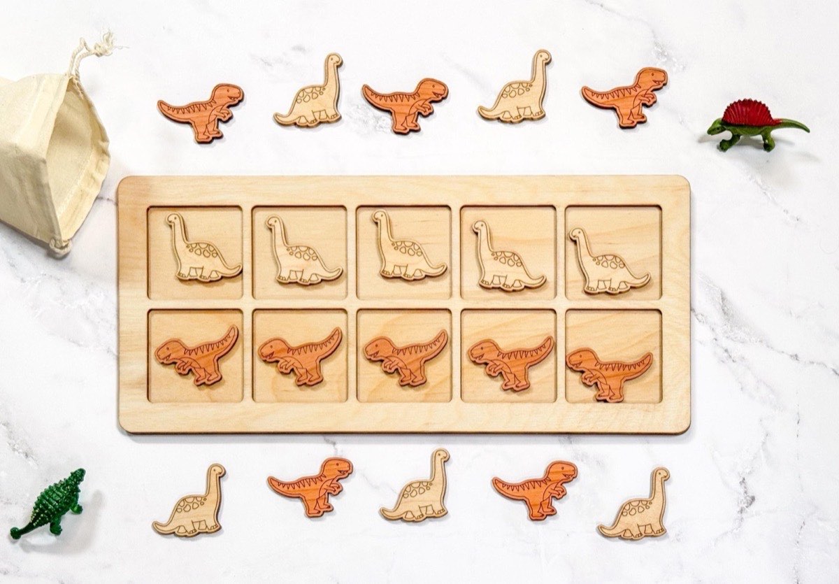 Non-personalized wooden ten frame in neutral Baltic birch, five light Brachiosaurus and five darker-toned T-rex counters in two rows, ringed by extra dinosaur pieces, toy figurines, and a natural muslin bag
