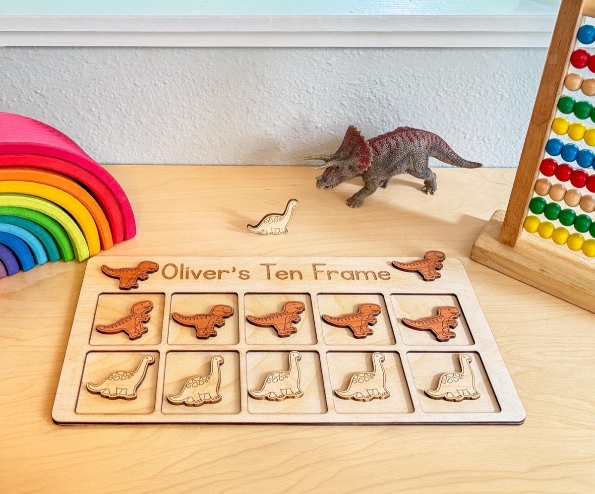 Dinosaur ten frame on a playroom desk with a toy Triceratops, rainbow stacker, and bead abacus, five T-rex counters in the top row and five Brachiosaurus counters below
