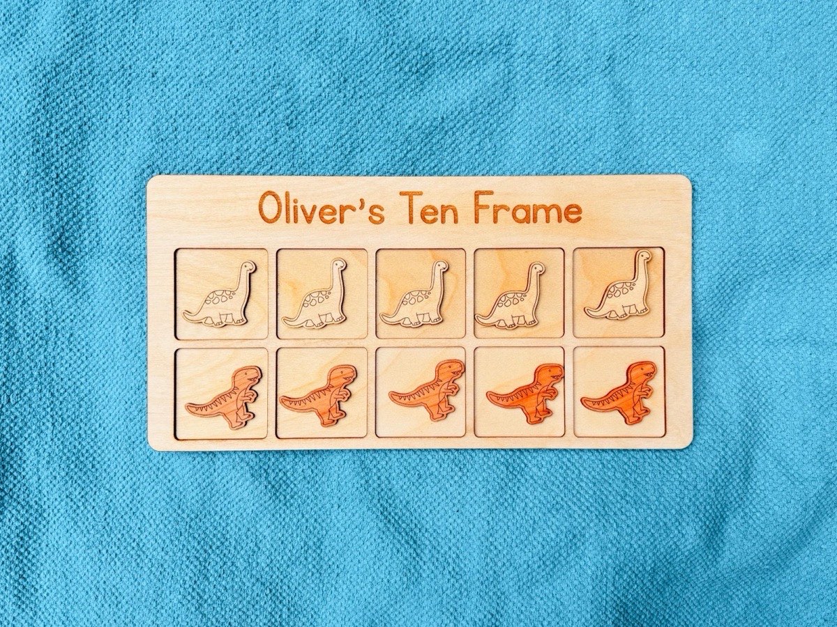 Overhead view of a personalized dinosaur ten frame on a teal floor mat, a row of five Brachiosaurus counters above a row of five T-rex counters in contrasting natural wood tones