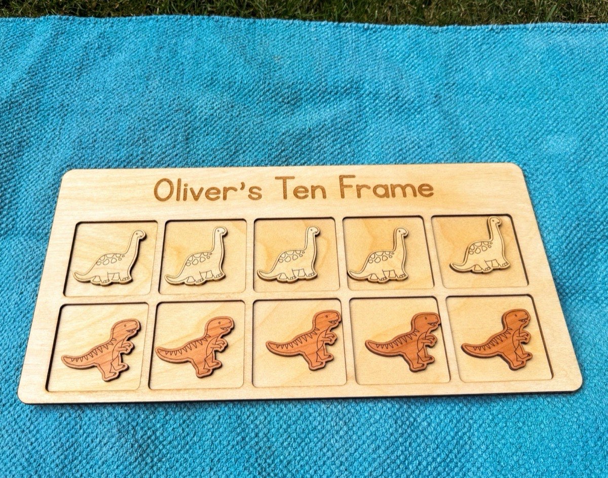 Overhead view of a personalized dinosaur ten frame on a teal floor mat with grass in the far background, a row of five Brachiosaurus counters above a row of five T-rex counters in contrasting natural wood tones