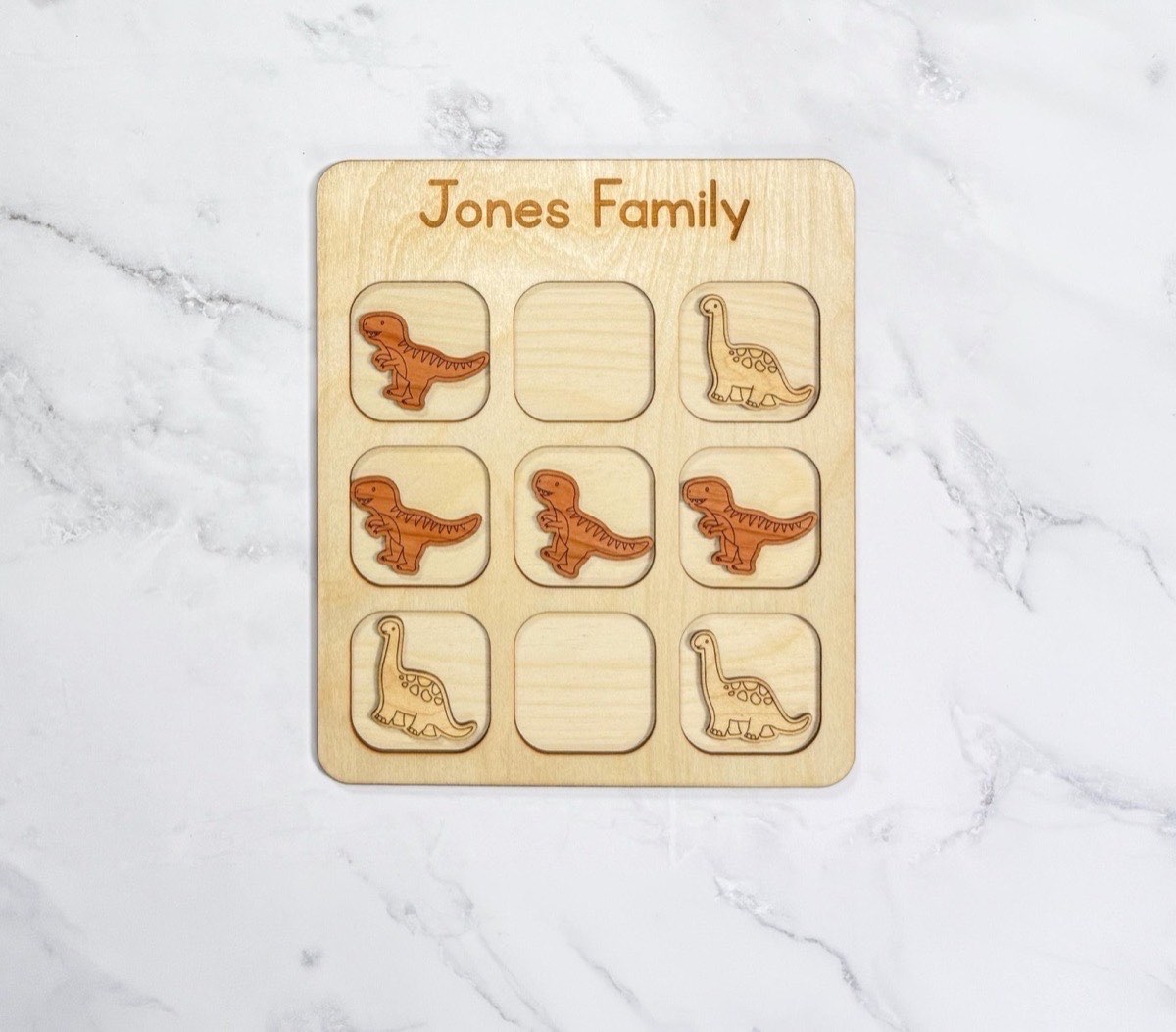 Personalized dinosaur tic tac toe board engraved 'Jones Family' on white marble, mid-game with darker T-rex pieces and lighter Brachiosaurus pieces on the rounded three-by-three grid