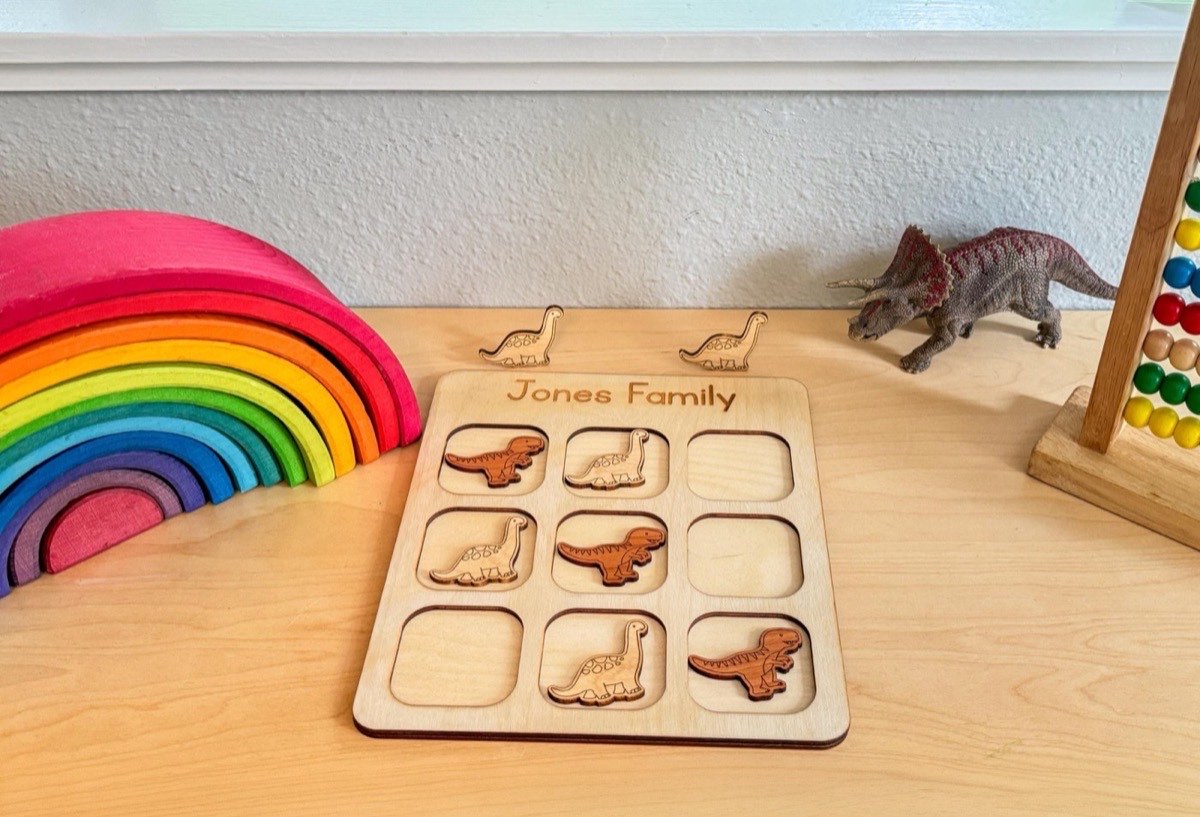Dinosaur tic tac toe on a playroom desk beside a rainbow stacker, toy Triceratops, and abacus, wooden T-rex and Brachiosaurus pieces mid-game with two extras above the board