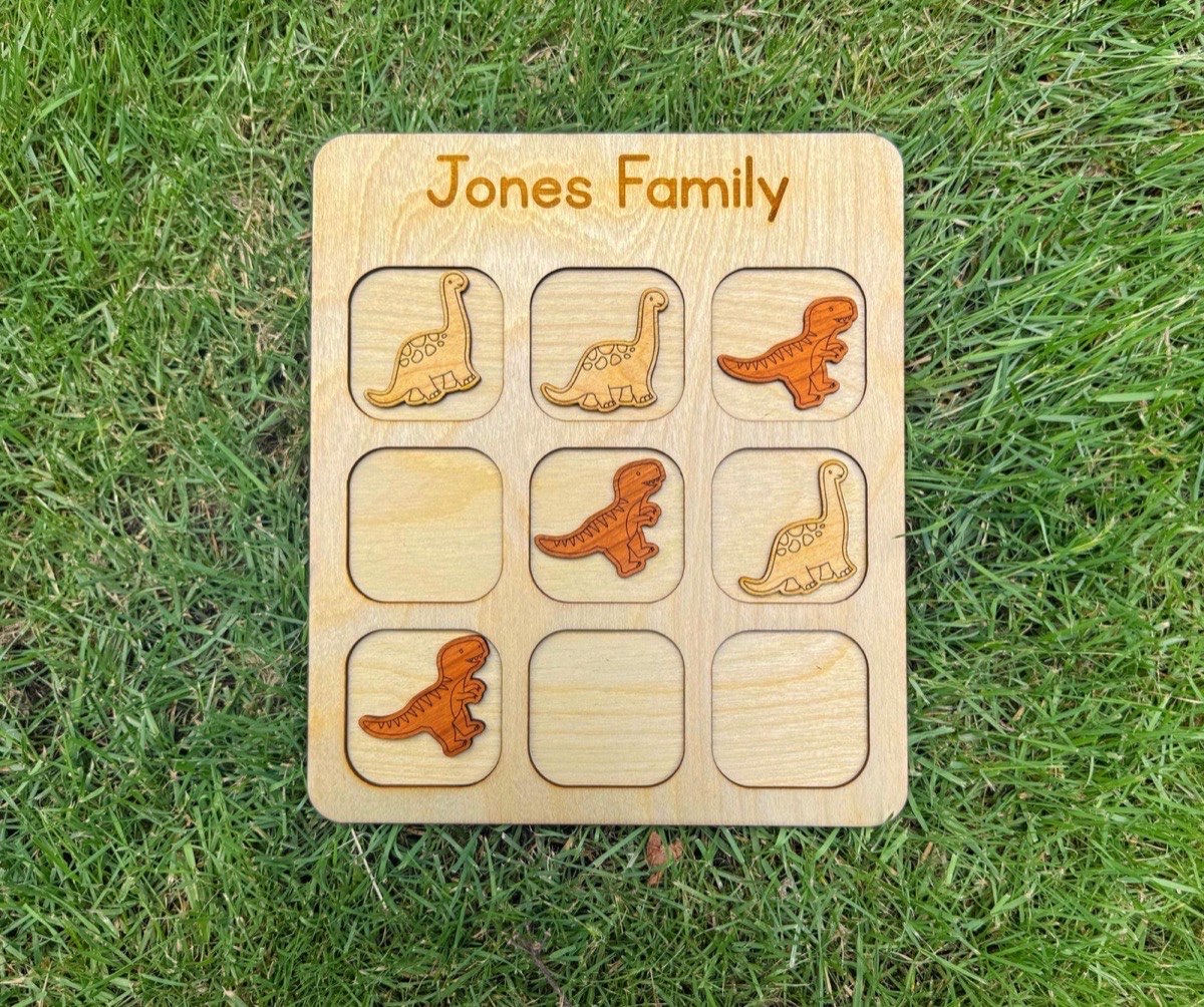 Close view of a wooden dinosaur tic tac toe game lying in grass, laser-engraved T-rex and Brachiosaurus pieces in contrasting natural wood tones on a board engraved 'Jones Family.'