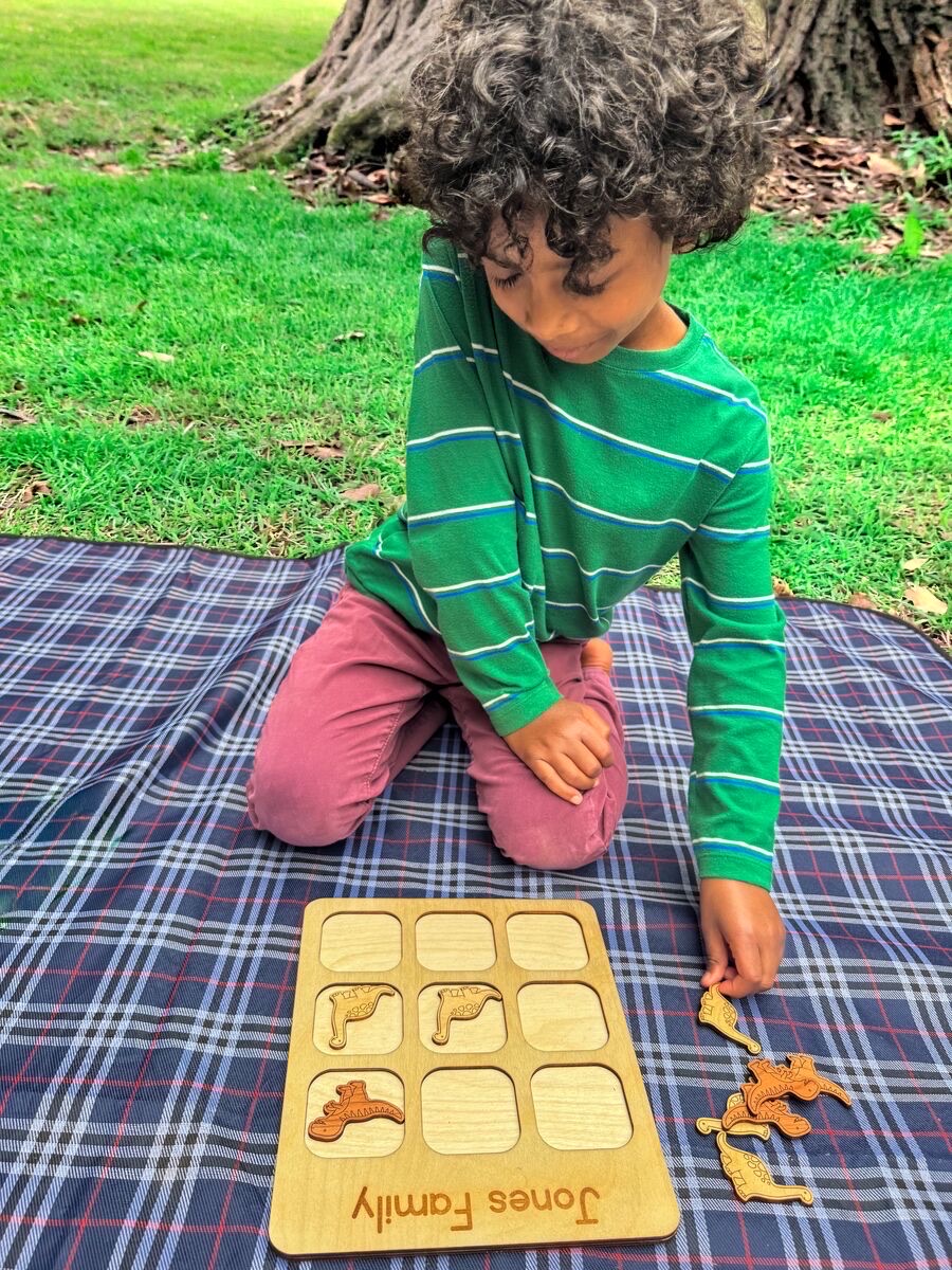 A child kneeling on a navy plaid blanket in grass places wooden dinosaur pieces on a tic tac toe board engraved 'Jones Family', two pale maple Brachiosaurus and one cherry T-rex on the grid with three pieces waiting beside it