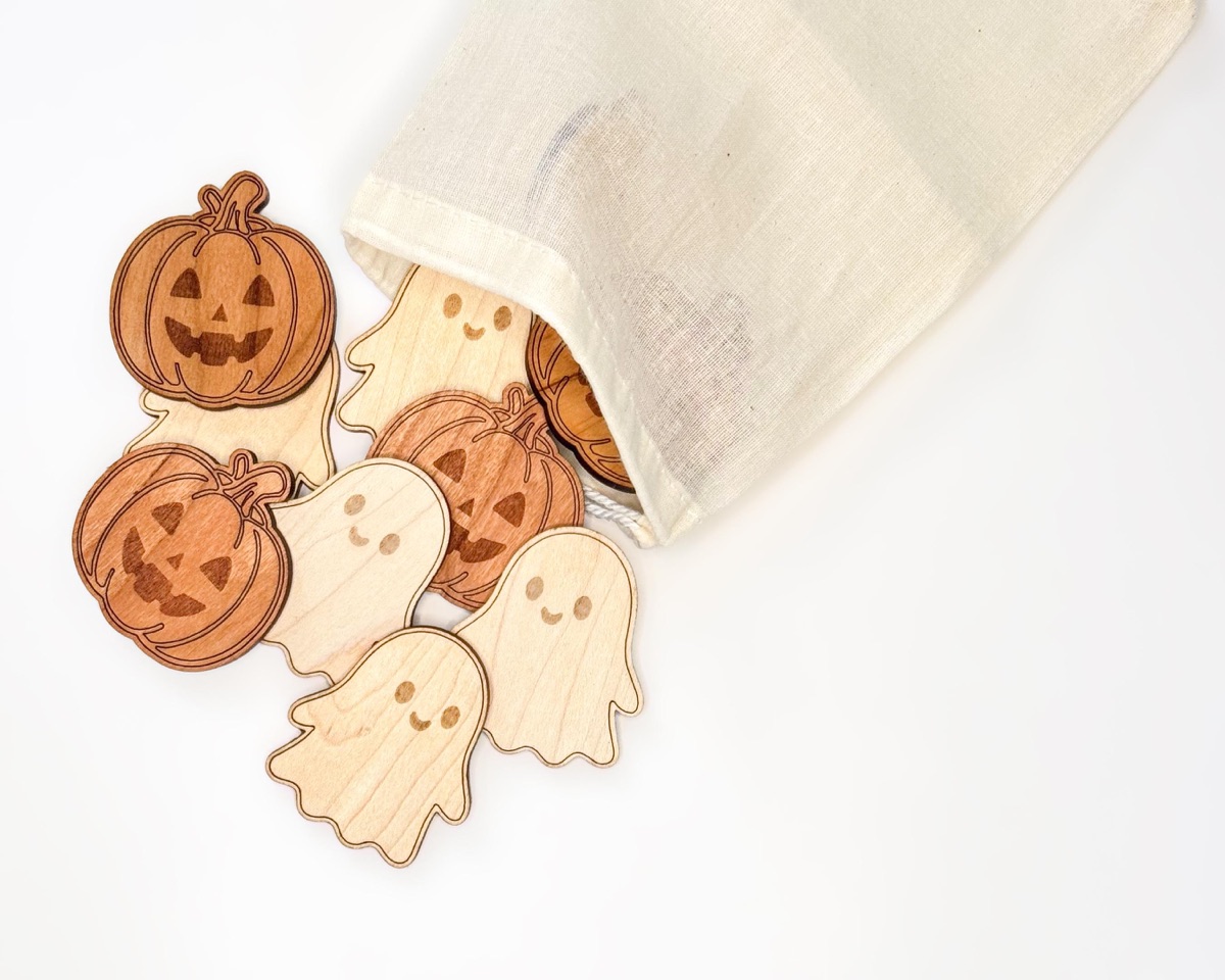 Maple ghost counters and cherry jack-o'-lantern counters spilling from a natural cotton drawstring bag onto a white surface, the two wood tones mixed together in a loose pile