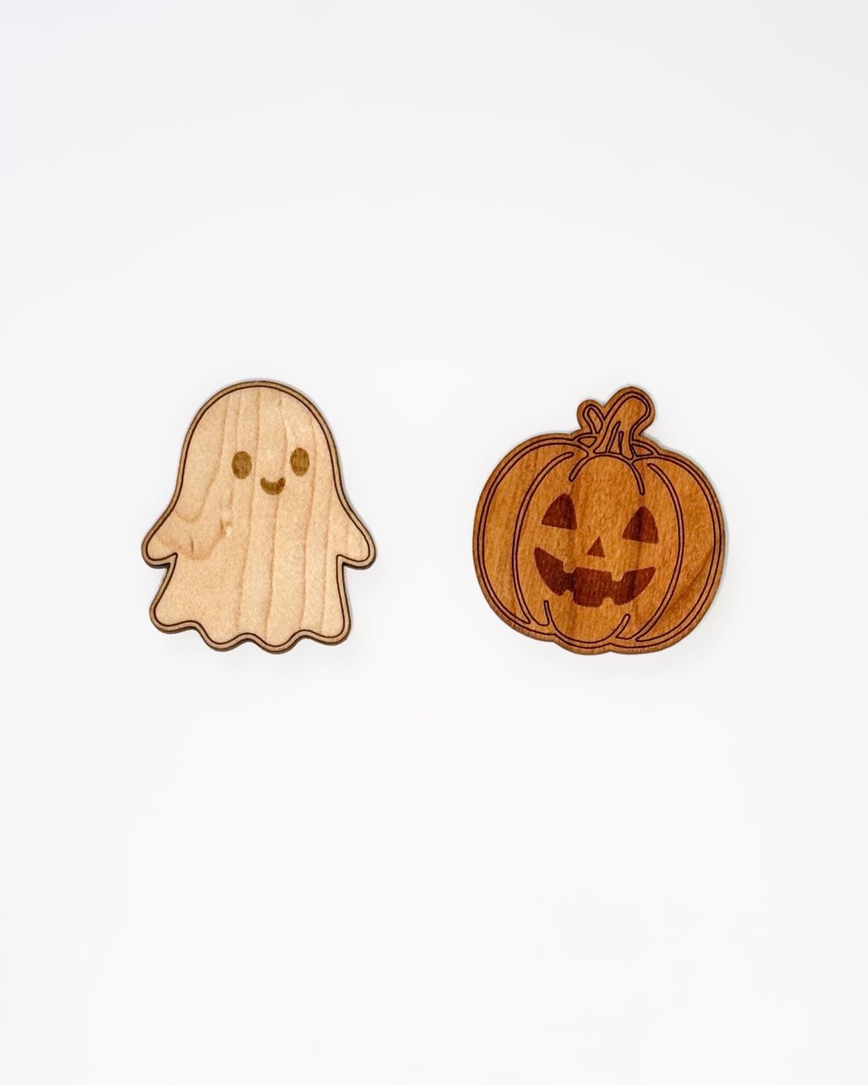 One maple ghost counter and one cherry jack-o'-lantern counter side by side on white, the pale ghost engraved with dot eyes and a smile, the reddish pumpkin with ribs, a curled stem, and a toothy face