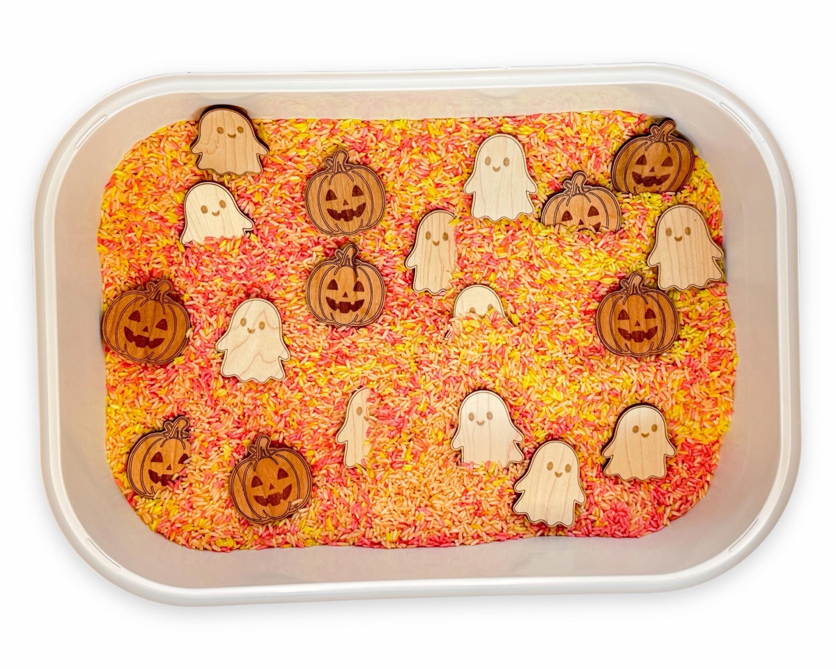 White rectangular tub of orange, pink, and yellow dyed rice holding eleven maple ghost counters and eight cherry jack-o'-lantern counters, seen from directly above on a white background