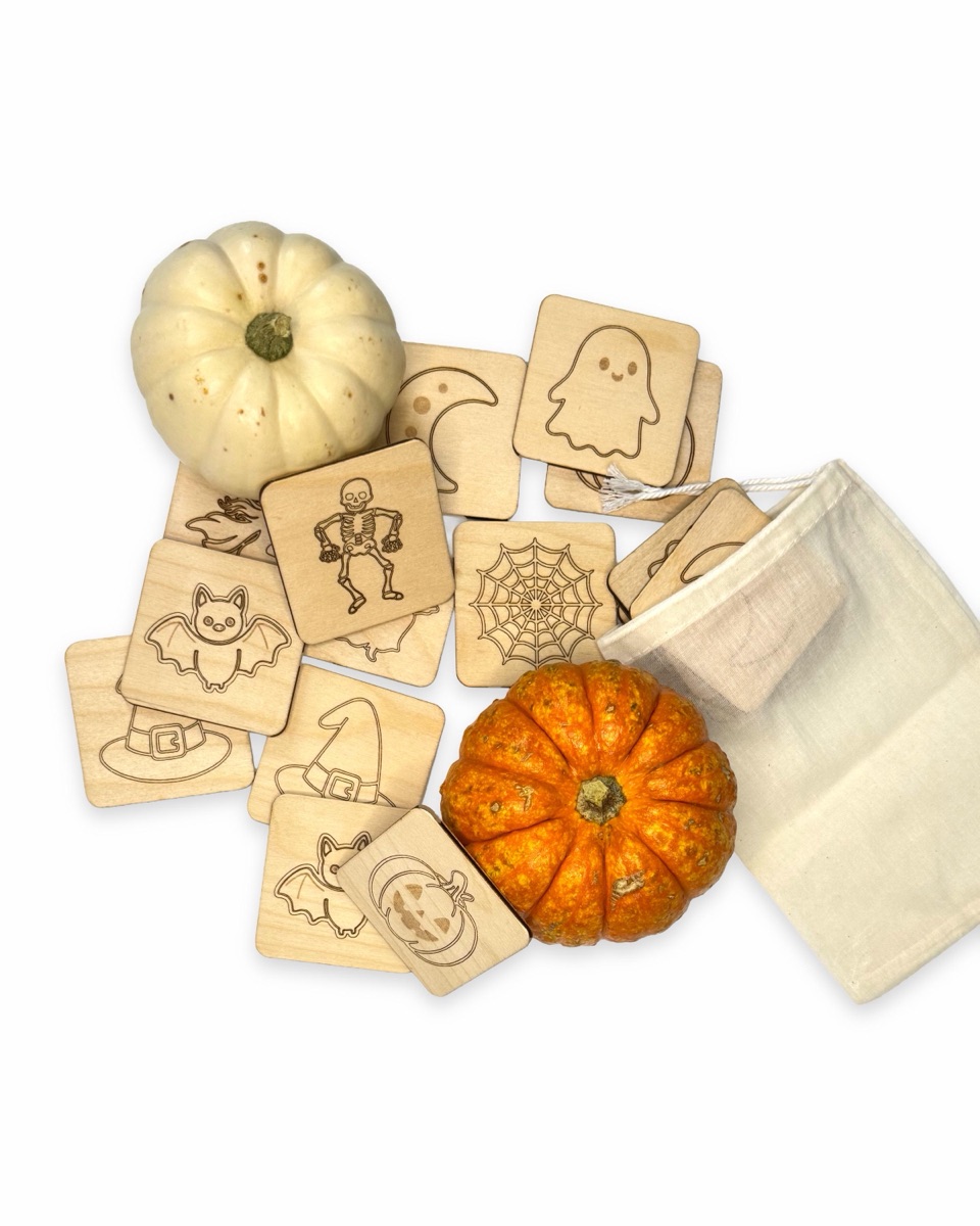Halloween memory tiles spread across a white surface with a small white pumpkin, an orange pumpkin, and a natural cotton drawstring bag, with the skeleton, spider web, ghost, and bat designs on top
