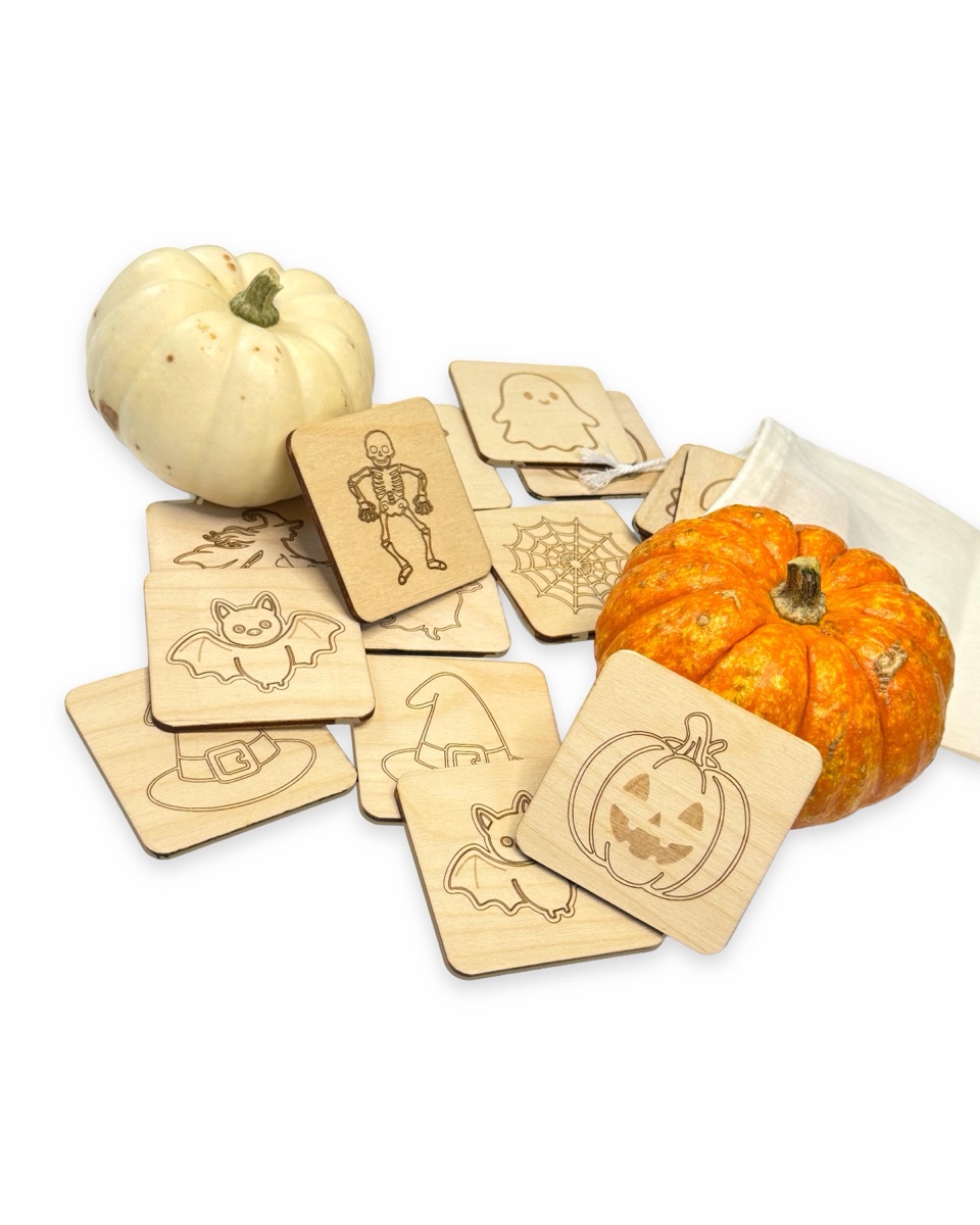 Halloween memory tiles piled at an angle beside a white pumpkin and an orange pumpkin, with the skeleton tile leaning on the white pumpkin and the jack-o'-lantern tile propped against the orange one