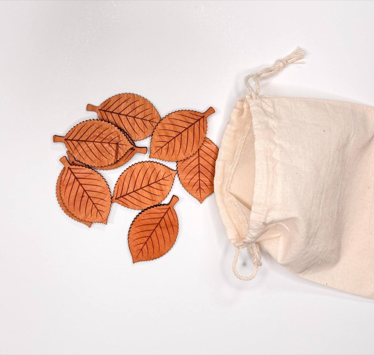 Seven solid cherry leaf counters with laser-engraved veins and serrated edges in warm reddish wood tones, shown beside an upright natural cotton drawstring pouch