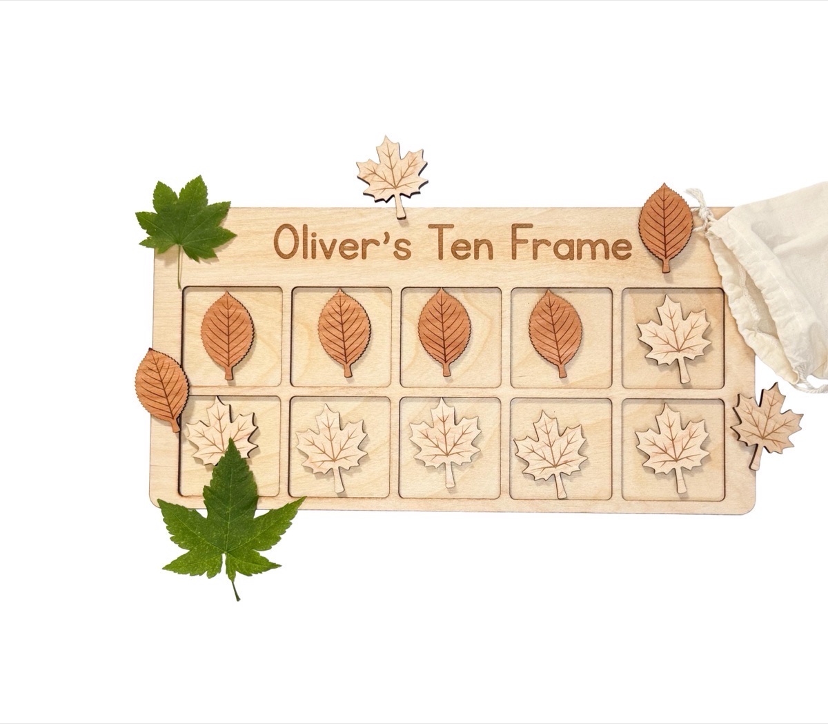 Baltic birch ten frame engraved 'Oliver's Ten Frame' on white, four cherry leaves and one maple leaf in the top row and five maple leaves below, loose leaves and a cotton bag around it beside two real green leaves