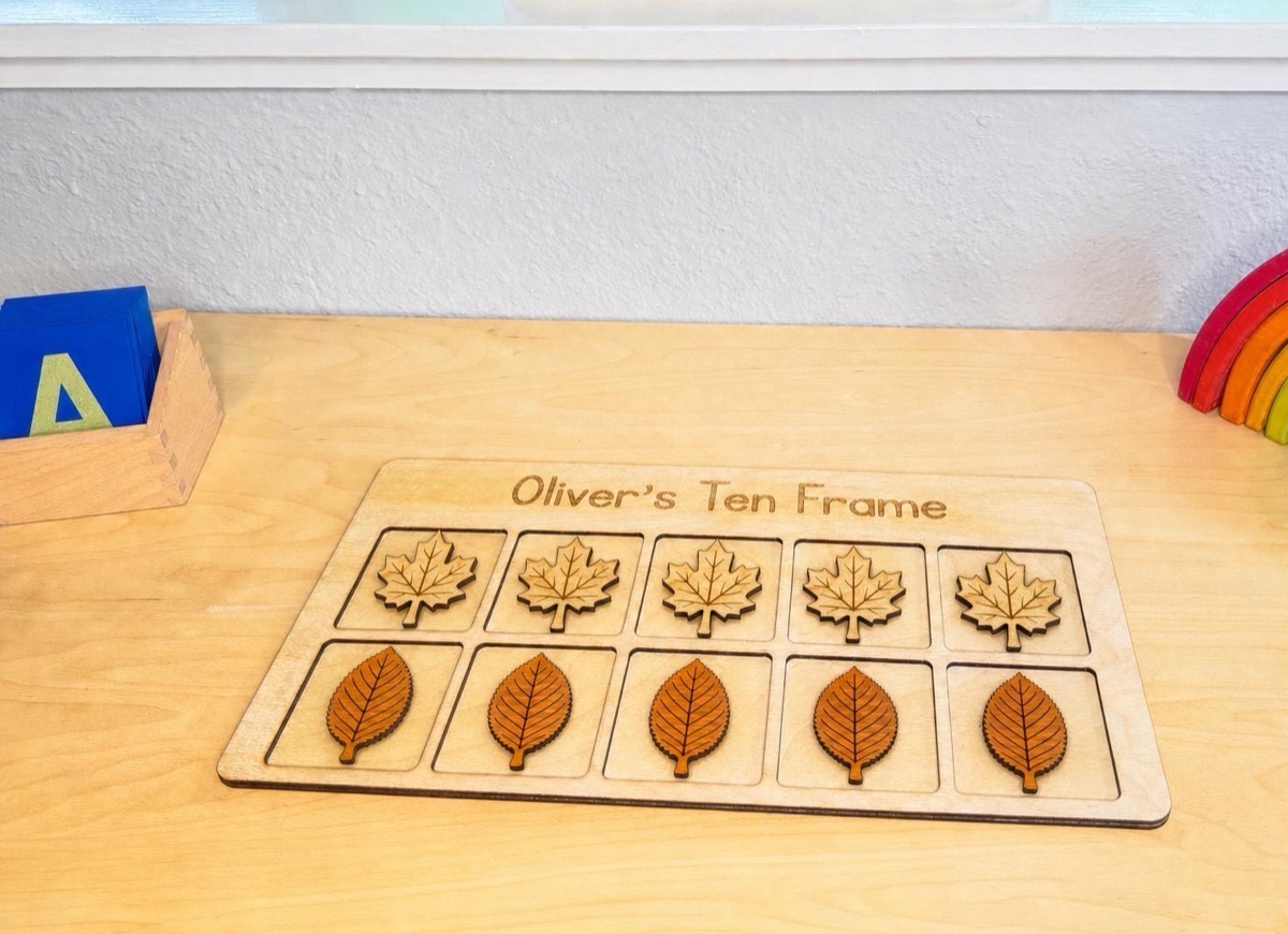 Leaf ten frame engraved 'Oliver's Ten Frame' on a wooden desk beside a box of blue sandpaper letters and a rainbow stacker, five maple leaves in the top row and five cherry leaves in the bottom row