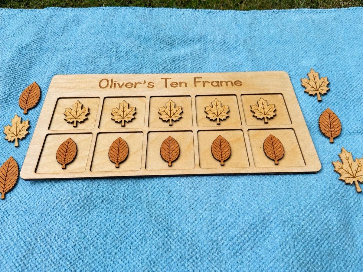 Leaf ten frame engraved 'Oliver's Ten Frame' on a blue blanket in grass, five maple leaves above five cherry leaves in the board with eight more leaves scattered on the fabric around it