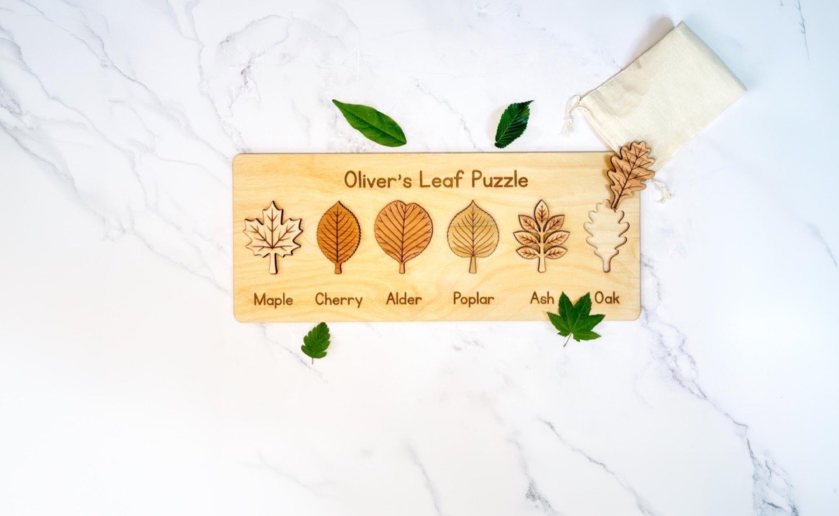 Personalized Montessori leaf puzzle engraved 'Oliver's Leaf Puzzle' with six labeled wooden leaves - maple, cherry, alder, poplar, ash, and oak - the oak piece resting on a muslin bag beside fresh green leaves on marble