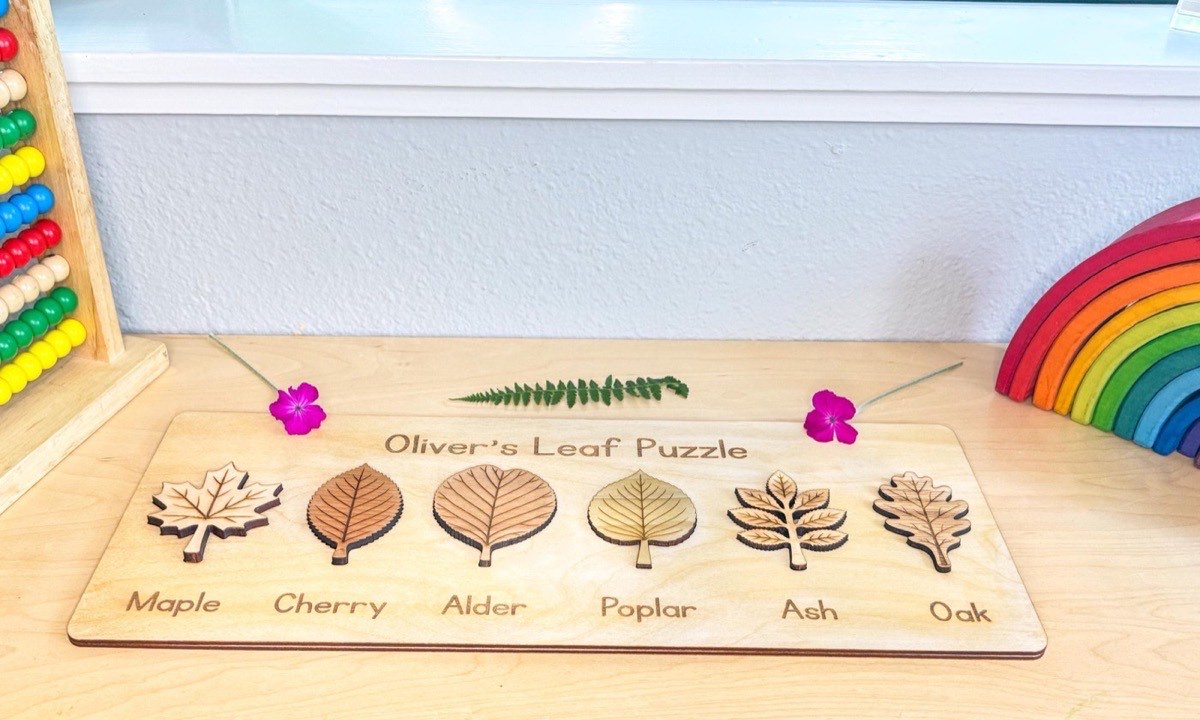 Personalized leaf puzzle on a playroom desk with a fern frond, pink blossoms, wooden rainbow stacker, and abacus, six labeled wooden leaves seated in the board. The wooden leaves are labeled - maple, cherry, alder, poplar, ash, and oak.  Each leaf is laser-engraved with realistic veins above its engraved name label