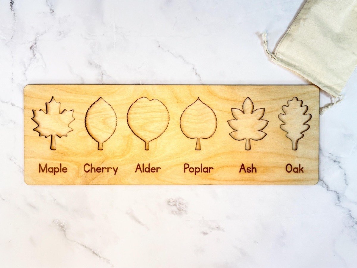 Empty leaf puzzle board showing six leaf-shaped recesses with engraved name labels - maple, cherry, alder, poplar, ash, oak - in neutral Baltic birch beside a muslin bag