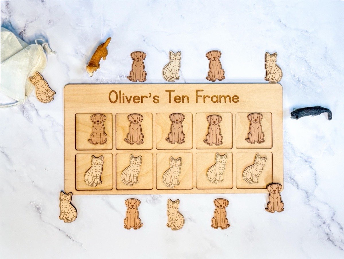 Personalized wooden ten frame engraved 'Oliver's Ten Frame' with five laser-engraved puppy counters and five tabby cat counters in place, extra pieces and toy animal figurines around the neutral Baltic birch board on white marble