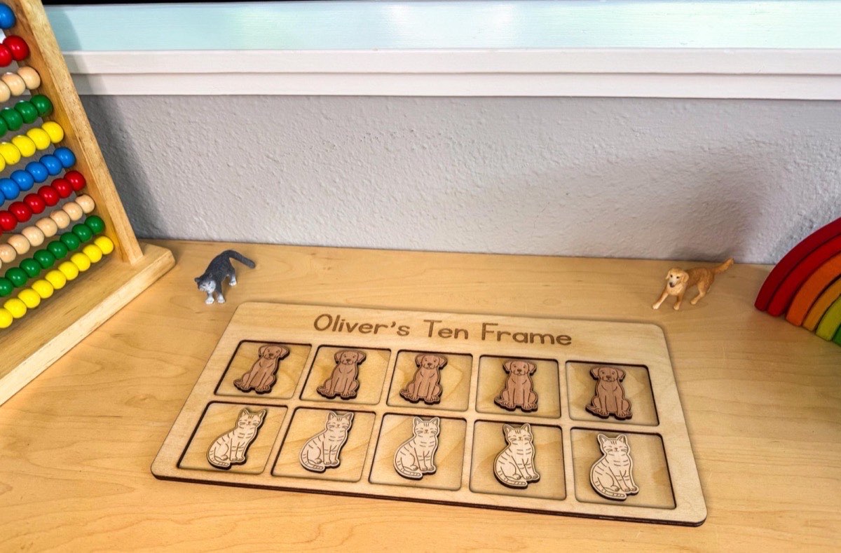 Cats and dogs ten frame on a playroom desk between a bead abacus, wooden rainbow stacker, and toy cat and dog figurines, ten neutral wooden counters seated in two rows of five