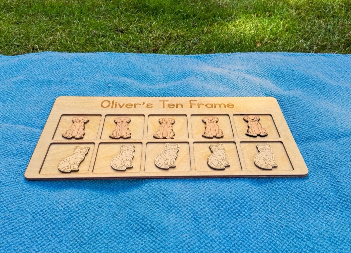 Ten frame counting board with dog and cat counters resting on a blue blanket in green grass, engraved with a child's name for personalized outdoor Montessori math practice
