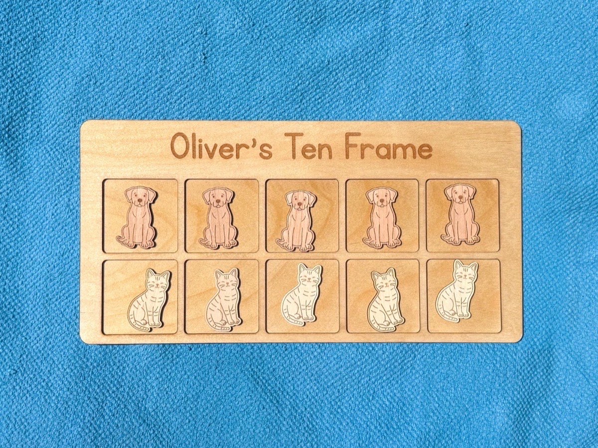 Wooden ten frame math board engraved 'Oliver's Ten Frame' on a bright blue floor mat, a row of five puppy counters above a row of five smiling cat counters, showing the laser-engraved detail
