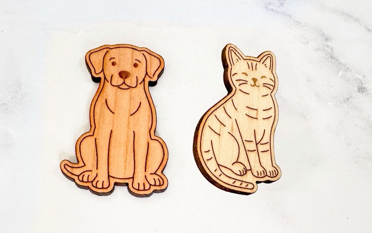 Close-up of two ten frame playing pieces: a sitting puppy in warm-toned wood and a smiling tabby cat in lighter neutral wood, both laser-engraved with rounded edges