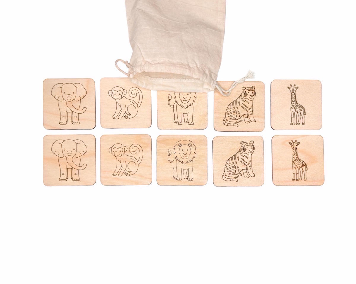 Ten wooden safari animal memory match tiles in two rows of five pairs, engraved with an elephant, monkey, lion, tiger, and giraffe, cut from pale birch with a natural cotton drawstring bag behind them