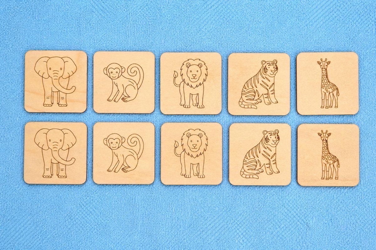 Complete ten-tile set laid out in two rows on a blue textured mat, showing all five matching pairs of elephant, monkey, lion, tiger, and giraffe engraved in fine outline on square wooden tiles with rounded corners