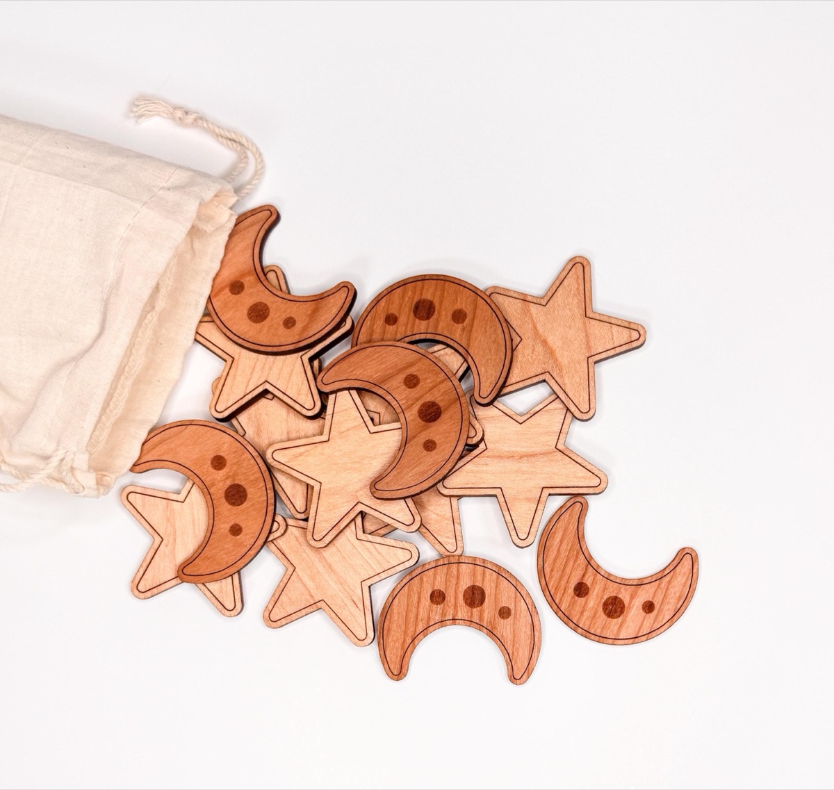 Wooden pale maple stars and warm cherry moon counters spilling from a cotton drawstring bag, laser-engraved moons and stars in undyed hardwood for Montessori counting, sorting, and fall sensory bin play