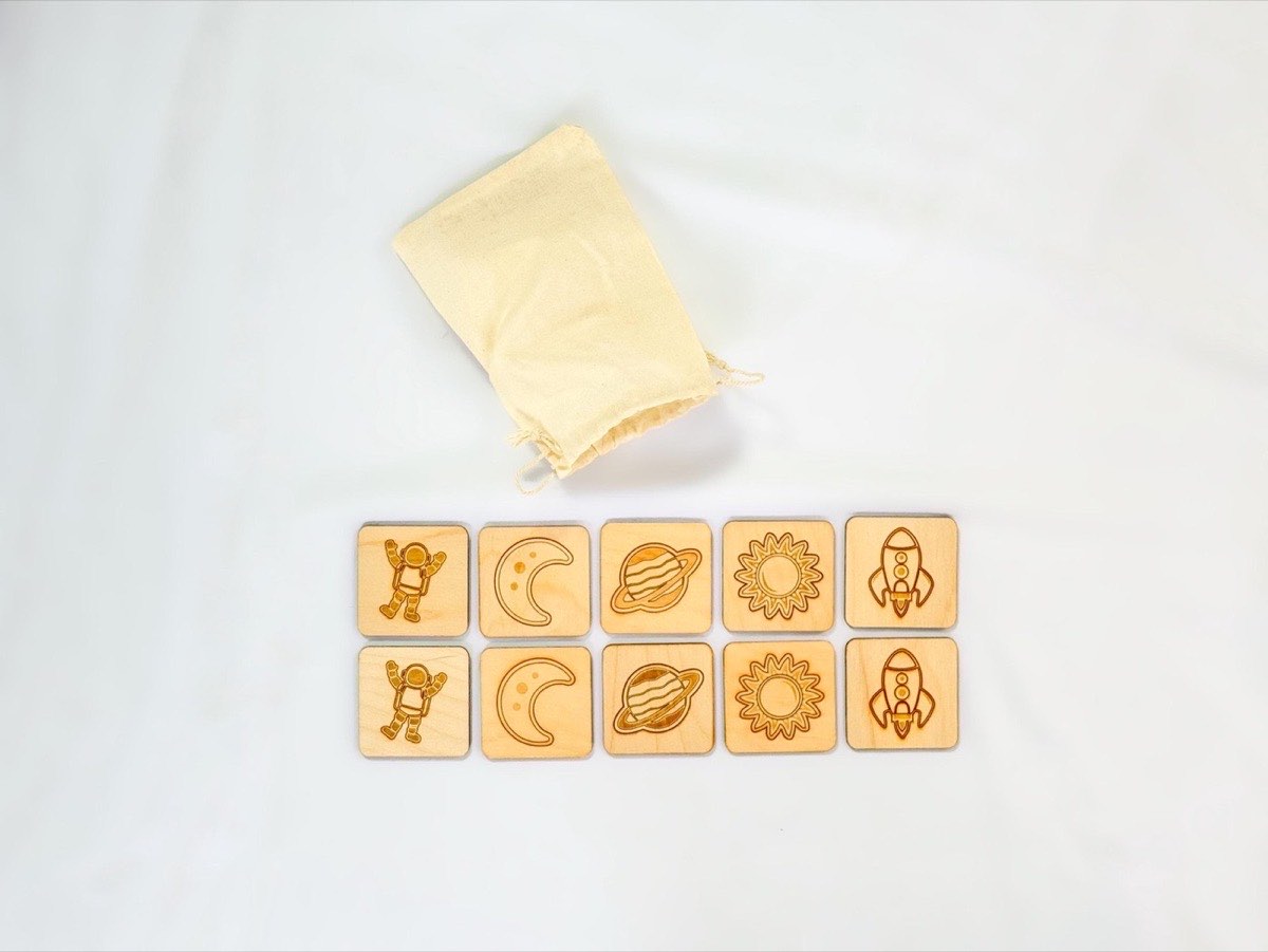 10-piece Montessori space memory match game, five matching pairs of astronaut, crescent moon, Saturn, sun, and rocket wooden tiles on Baltic Birch.  Displayed on white fabric background