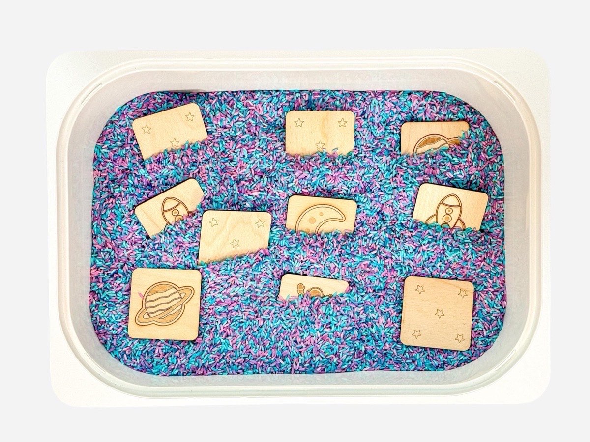 Ten double-sided natural colored wooden space memory tiles partially buried in purple and teal rainbow rice sensory bin