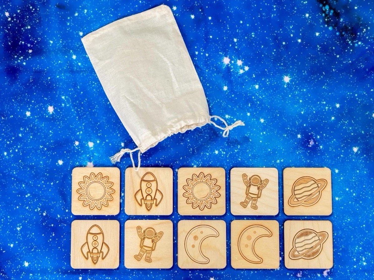 Ten-piece neutral-colored wooden space memory match set arranged on starry night backdrop, featuring five pairs of laser-engraved astronaut, moon, planet, sun, rocket tiles